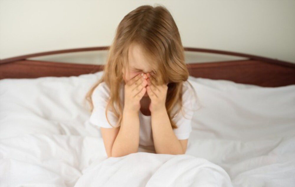 A little girl sits and sneezes on the bed among the blankets and pillows. May be due to dust mite allergy symptoms.