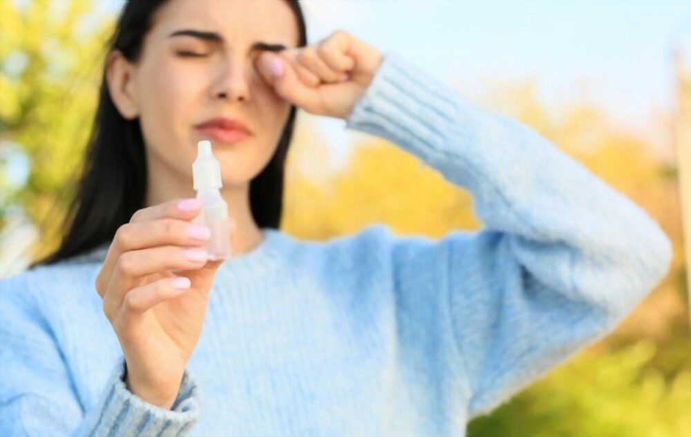Young woman with eye drops for allergies in hand standing outside and itching her eye.